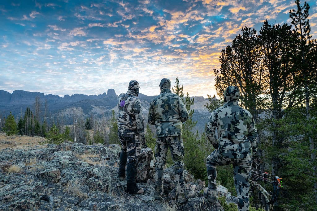 Three elk hunters facing a canyon with a sunset in the distance