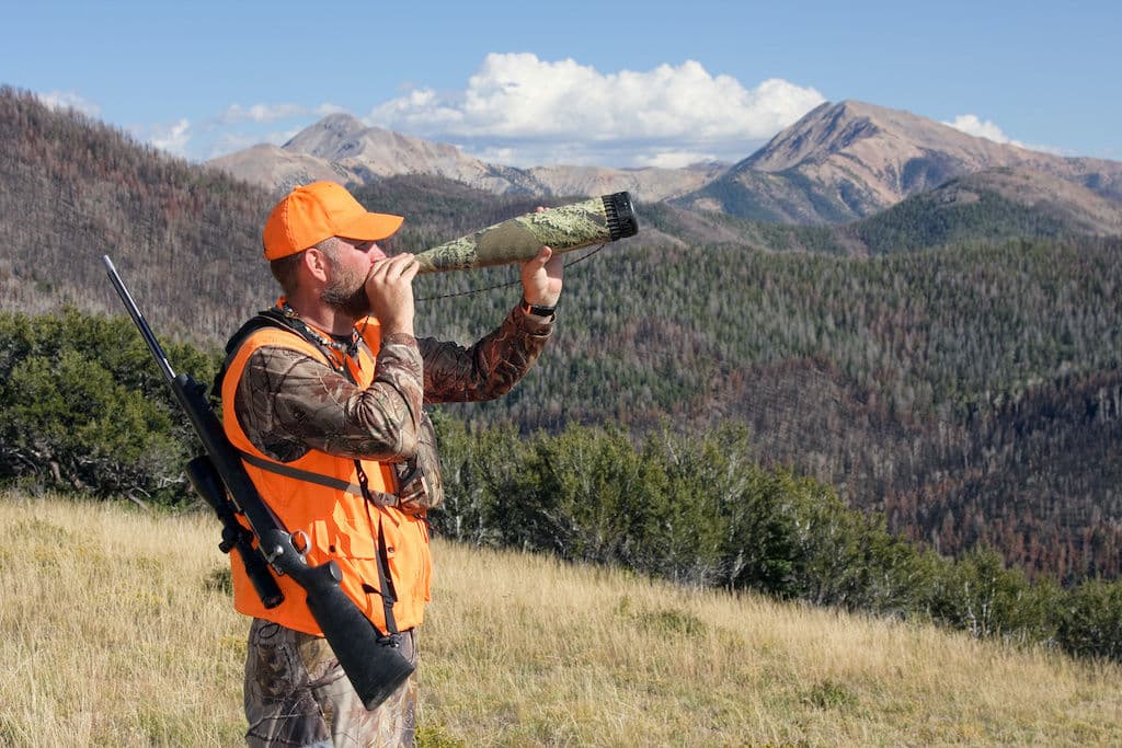 A man dressed in camo and orange blowing into an elk bugle tube