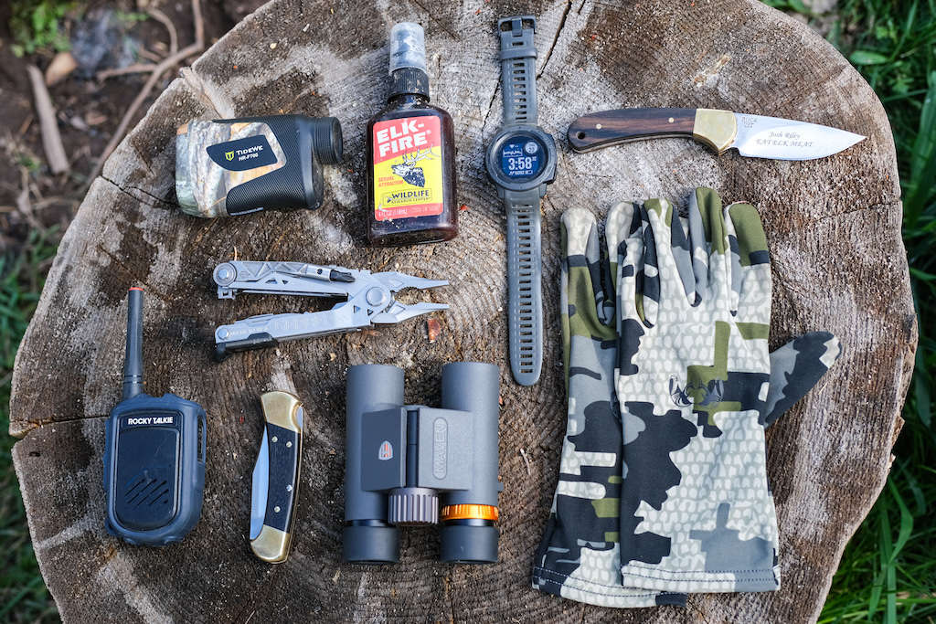 Rangefinder, elk pee, hunting watch, skinning knives, camo gloves, binoculars, Rocky Talkie, and a multi-tool laid out across a log