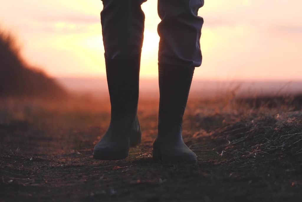 An elk and deer hunter with a pair of waterproof rubber boots walks down a dirt trail as the sun sets in the background