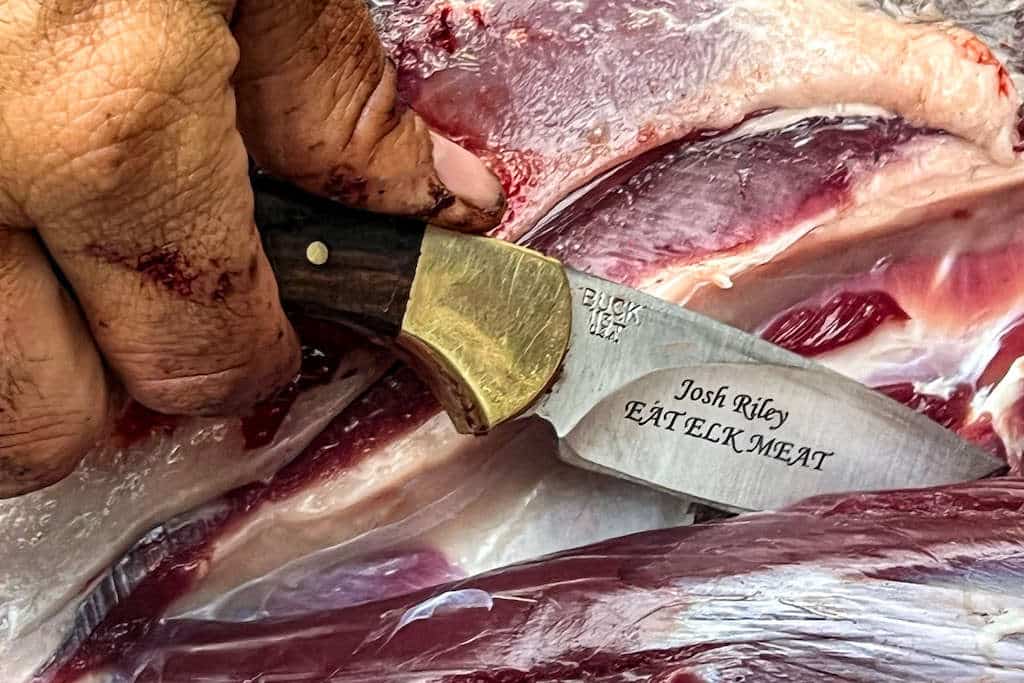 Hunter with a skinning knife reading "Josh Riley EAT ELK MEAT" field dressing a mule deer