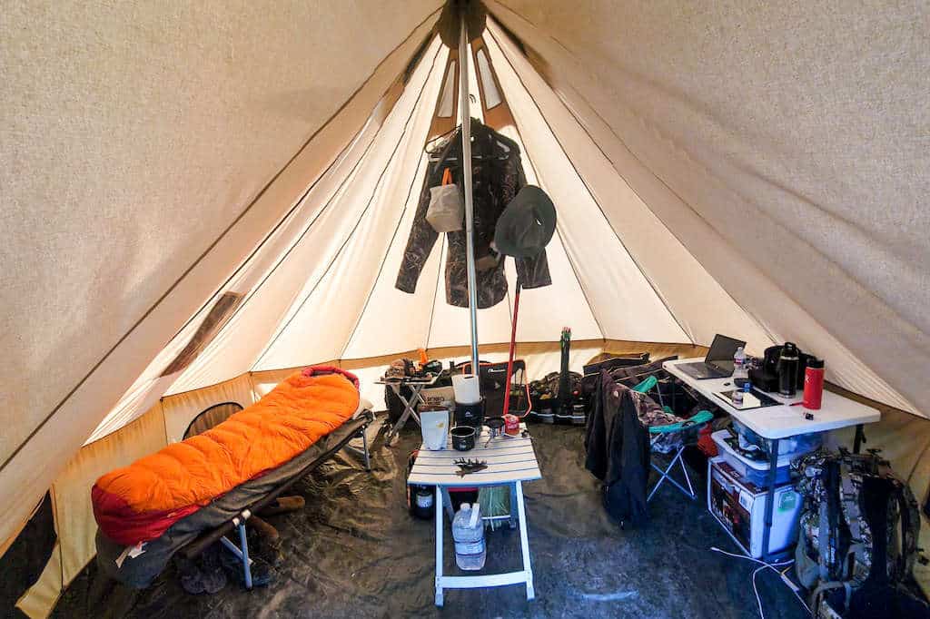 Inside of a hunting shelter with a cot, sleeping bag, tables, chair, coputer, and clothing