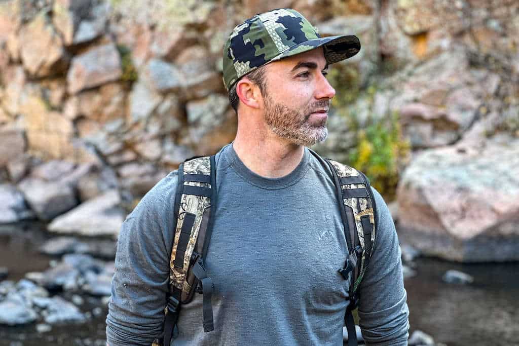 Hunter wearing a grey shirt, camo hat, and camo backpack looking towards the mountains