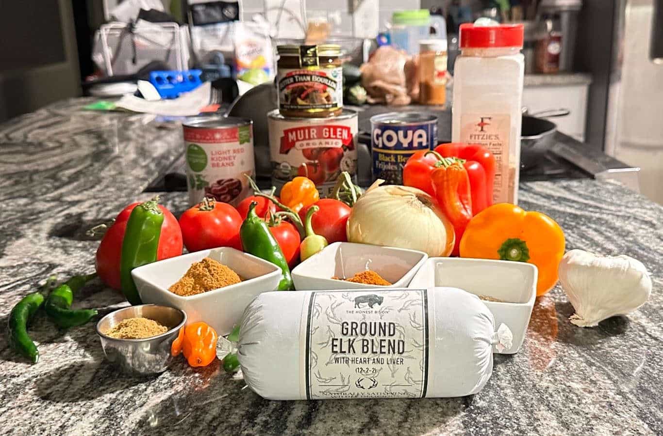 Ground elk meat, spices, tomatoes, various vegetables, cans of beans, seasoning, oil on a granite countertop