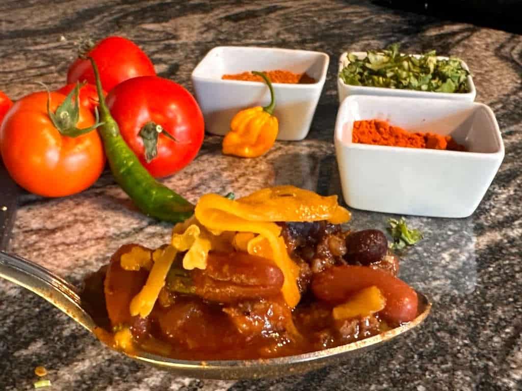 Spoonful of elk chili with bowls of spices and vegetables in the background