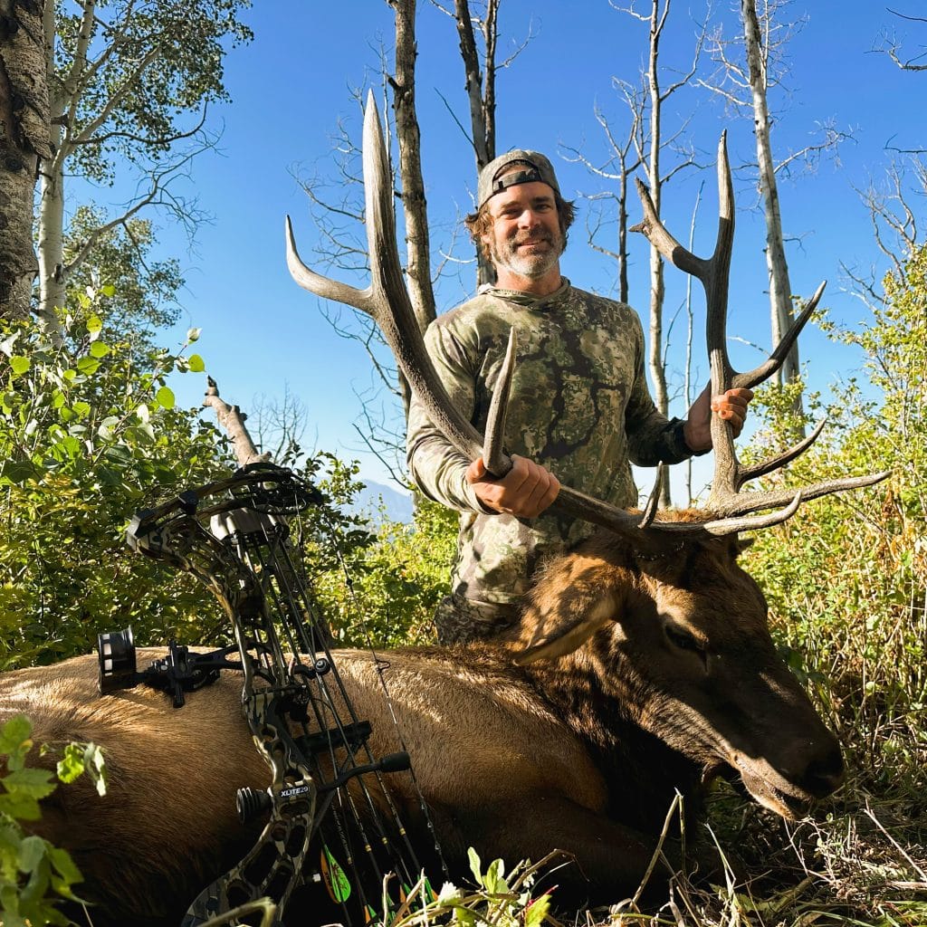 A hunter holding up an elk after harvesting it with an xpedition xlite mossey oak bottom land, with mechanical broadhead