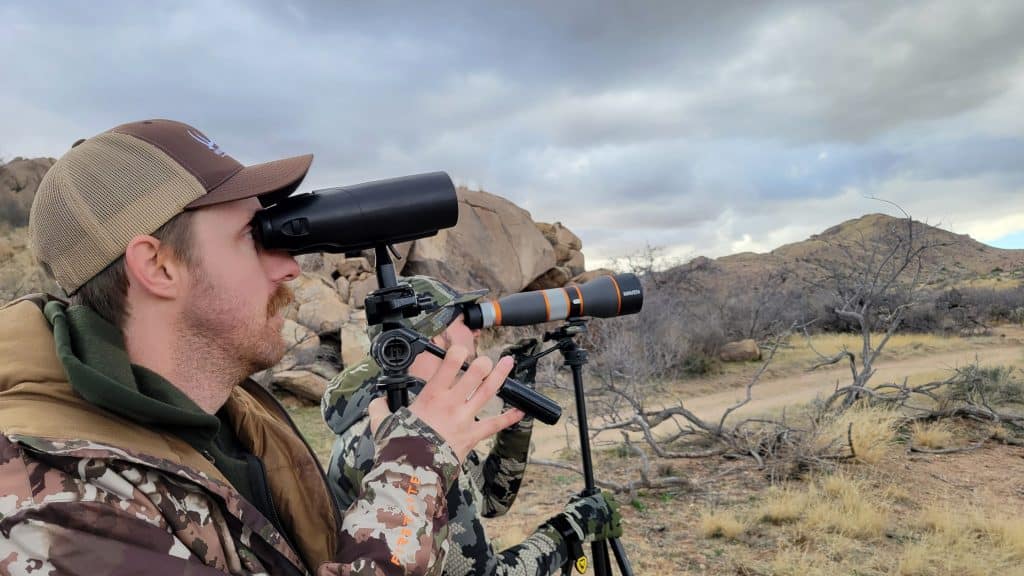 Two hunters glassing high desert country, one with binoculars and one behind a Maven spotting scope on a tripod