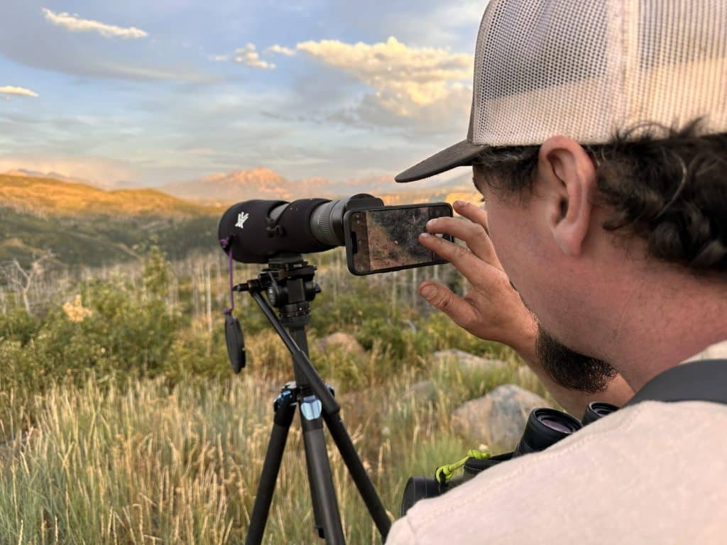 Hunter seated on a rock ledge with a Vortex spotting scope glassing a snow-capped Colorado mountain basin