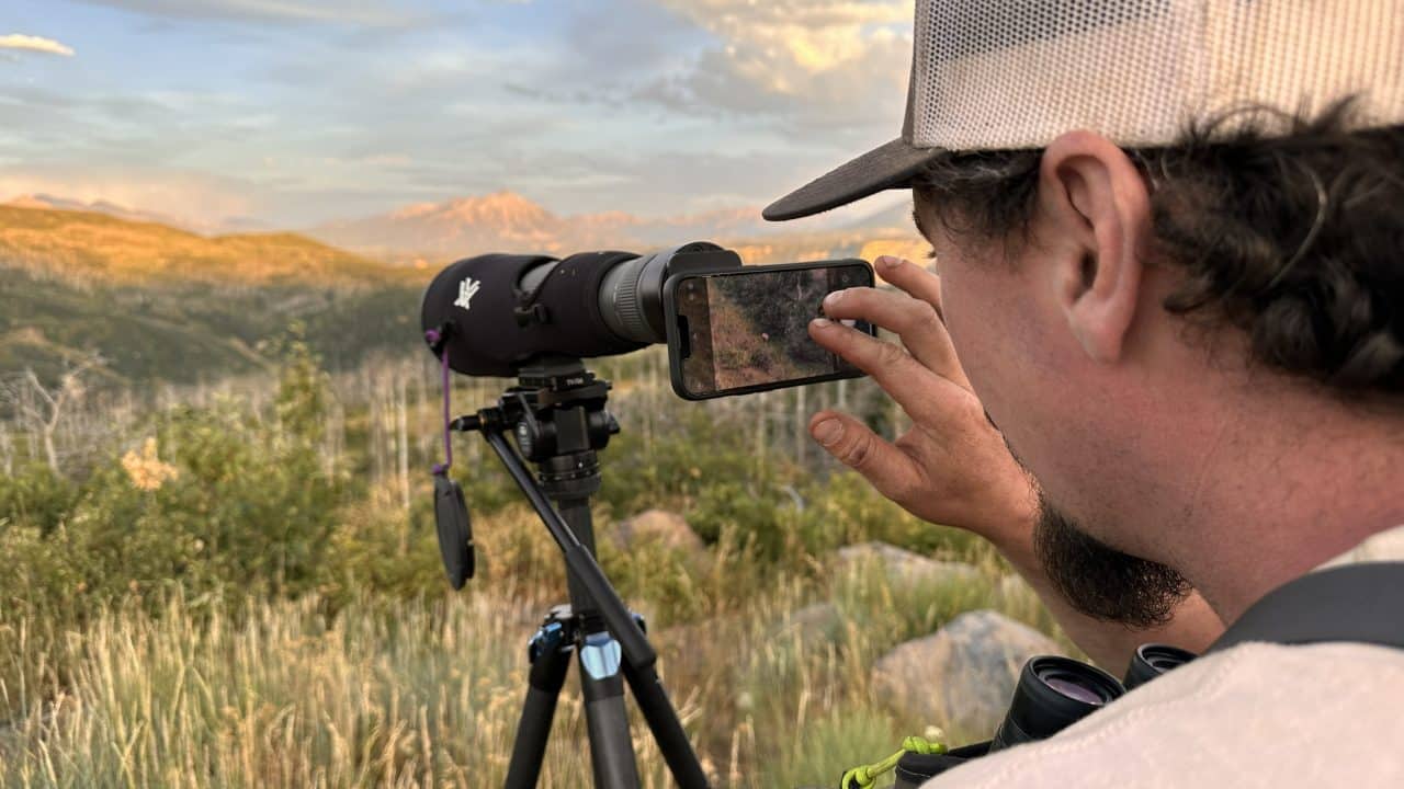 Hunter seated on a rock ledge with a Vortex spotting scope glassing a snow-capped Colorado mountain basin