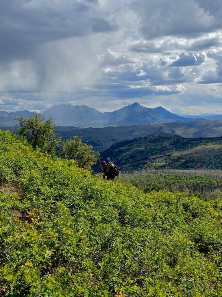 Bowhunter with a compound bow on his pack hiking through oakbrush as a rainstorm sweeps across the Colorado high country