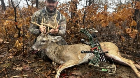 Bowhunter kneeling behind a whitetail buck with a compound bow after a broadhead pass-through in wet oak timber
